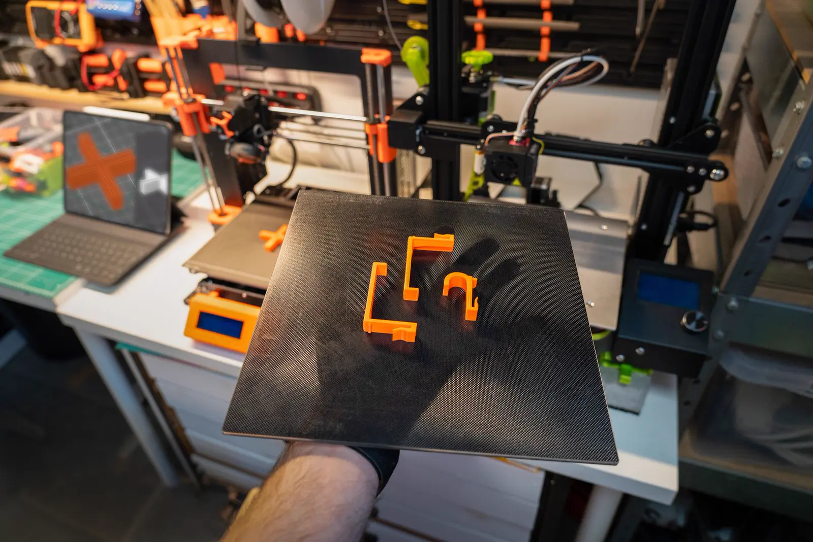 Plateau d’imprimante 3D tenu à la main, avec trois pièces orange fraîchement imprimées, devant des imprimantes