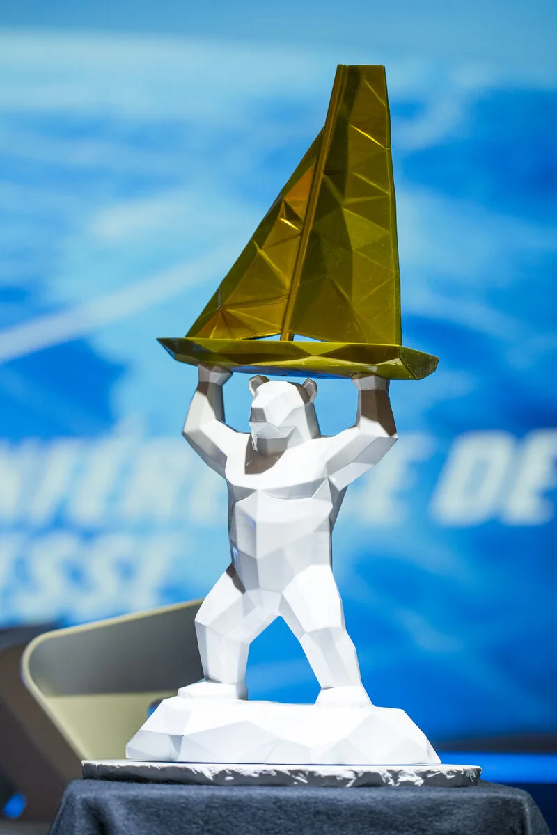 Trophée représentant un ours blanc à facettes qui soulève un voilier doré, présenté sur une table lors d’une conférence